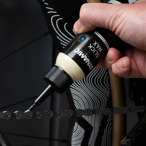 Hand applying lubricant to a bicycle chain with a bottle labeled 'Dinamic'.
